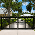 Black aluminum swing gate with X-brace design set between white stucco pillar columns with lantern lighting, opening to a paved driveway flanked by manicured tropical landscaping and a Florida estate home — residential gate installation in Sarasota County.
