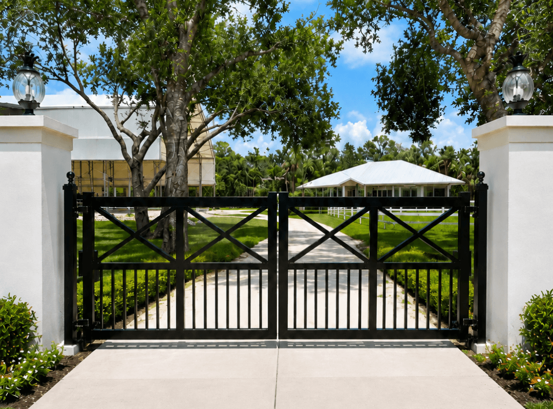 Black aluminum swing gate with X-brace design set between white stucco pillar columns with lantern lighting, opening to a paved driveway flanked by manicured tropical landscaping and a Florida estate home — residential gate installation in Sarasota County.